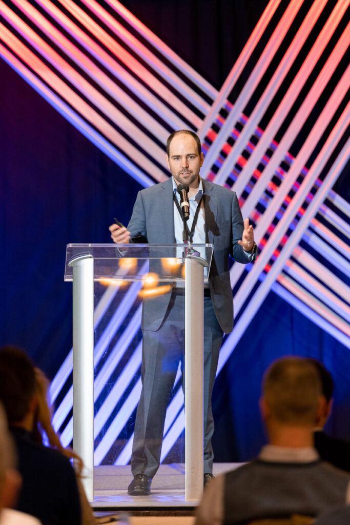 A confident speaker addresses an audience from behind a clear podium, against a dynamic backdrop of converging neon light lines.