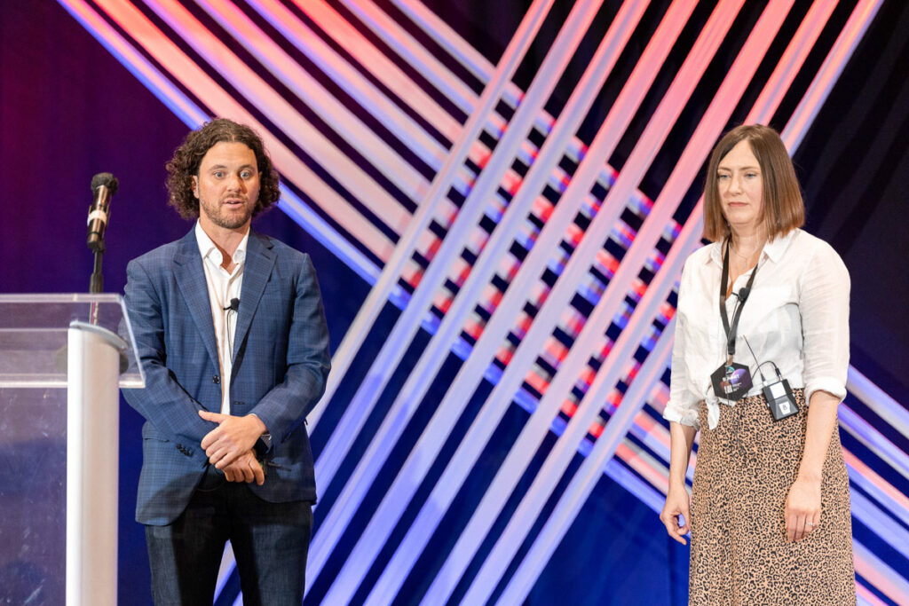 Two speakers at a conference stand in front of a colorful lit stage backdrop, with one gesturing expressively as they address the audience and the other looking on attentively.