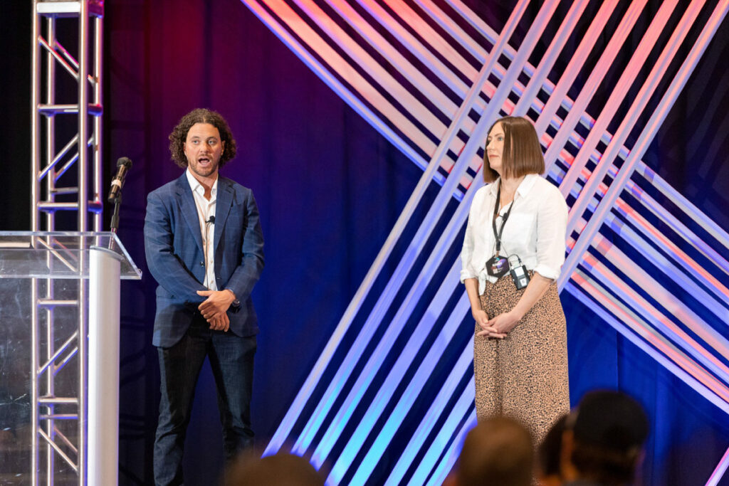 Two presenters at a conference, with one speaker actively engaging the audience while the other waits her turn by the podium against a backdrop of colorful stage lighting.