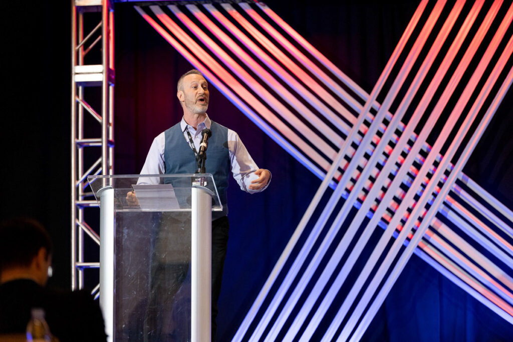 A man delivering a speech at a podium with dynamic lighting design in the background.