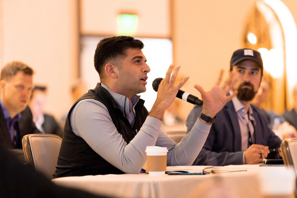 A man in a vest gesticulating while speaking at a conference, with other participants listening attentively.