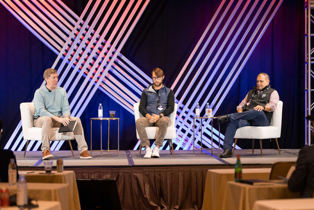 Three individuals participating in a panel discussion at a conference with a dynamic geometric background on the stage.