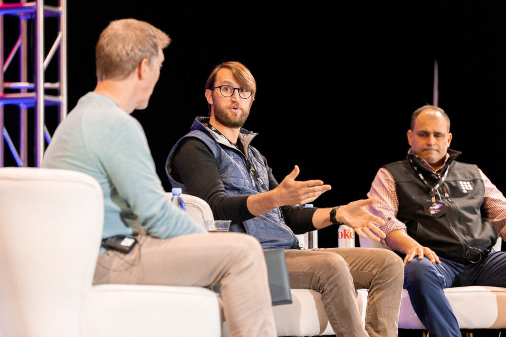 Three individuals engaged in a panel discussion at an event, with one of them gesturing while speaking, another attentively listening, and the third awaiting his turn to contribute to the conversation.