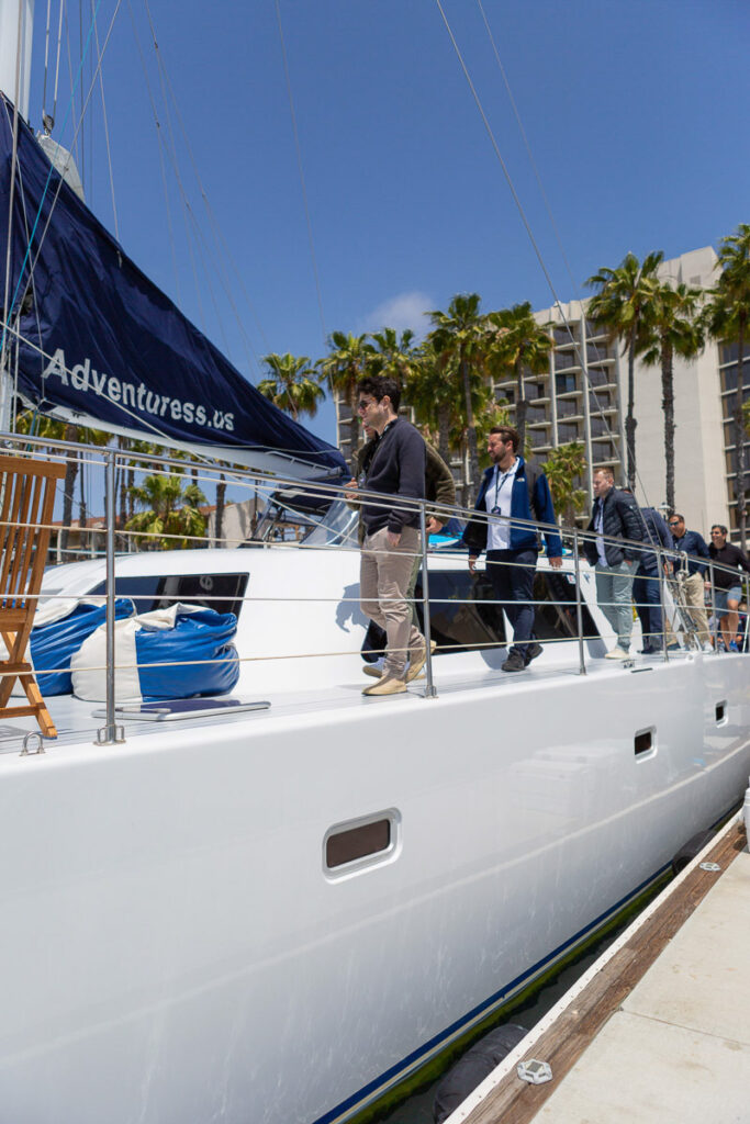 Boarding a sleek white sailboat on a sunny day at the marina, with palm trees and a clear blue sky in the background.