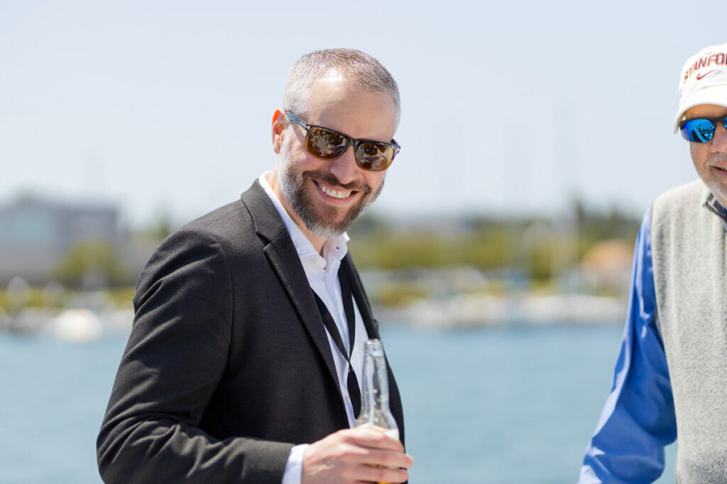 A man in sunglasses and a smart blazer smiling and holding a bottle, with clear blue skies and water in the background, enjoying a sunny day outdoors.