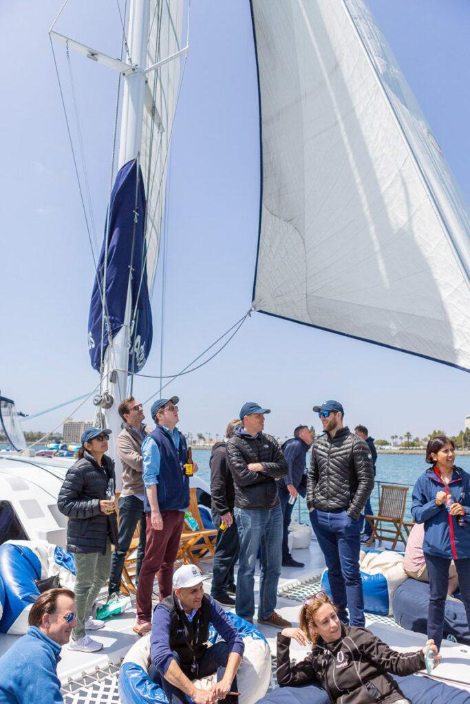 A group of people enjoying a sunny day on a sailboat, with some guests relaxing while others stand and converse, all surrounded by crisp white sails and a clear blue sky.