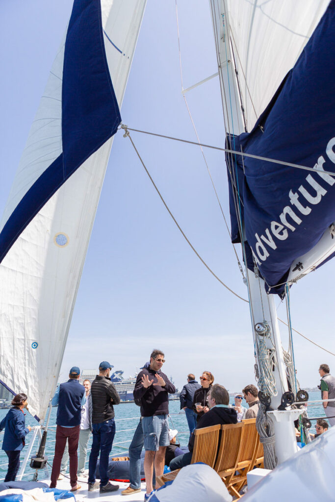 A group of people enjoying a sunny day on a sailboat with white and blue sails billowing in the wind against a clear blue sky.