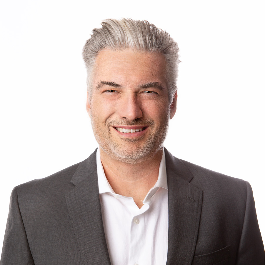 Portrait of John Mcleod, distinguished man with salt-and-pepper hair wearing a dark suit and a friendly smile against a white background.