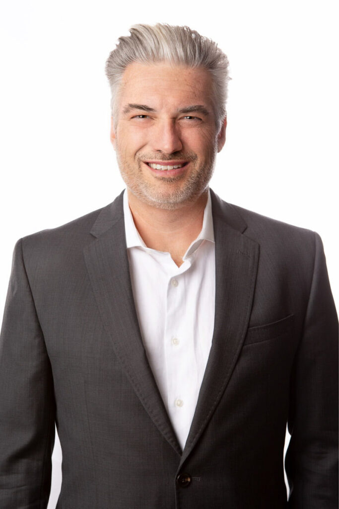 Portrait of John Mcleod, distinguished man with salt-and-pepper hair wearing a dark suit and a friendly smile against a white background.