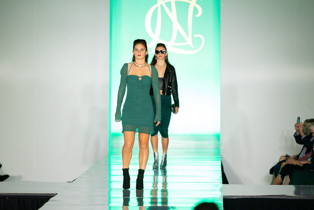 Two models confidently strutting down the fashion runway, with one leading in a green dress and the other following in a black ensemble, against the backdrop of a brightly lit designer logo.