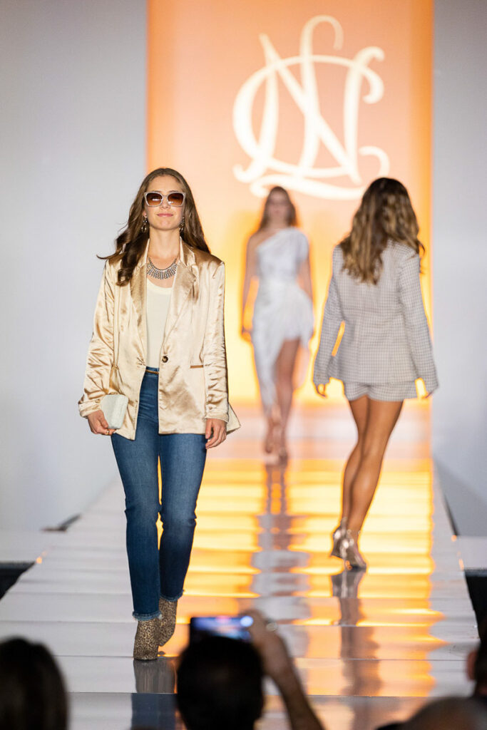 A confident model struts down the runway in a chic gold jacket and jeans combo, while another one follows her in the backdrop, both showcasing contemporary fashion designs.