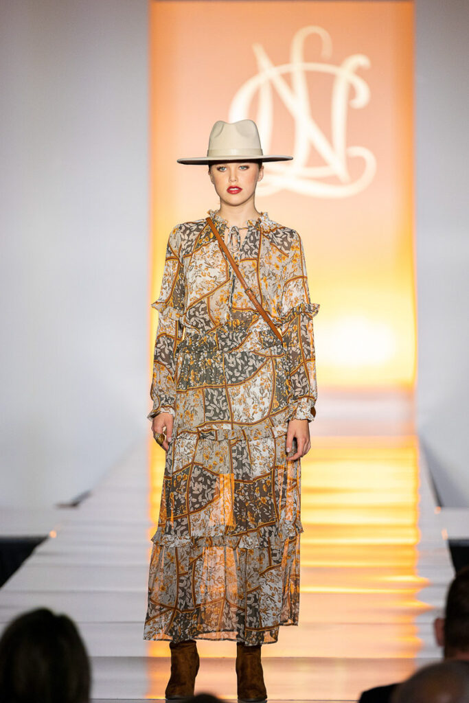 A model walks down the runway in a chic bohemian ensemble featuring a patterned long dress, accessorized with a crossbody bag and a wide-brimmed hat, against the warm glow.