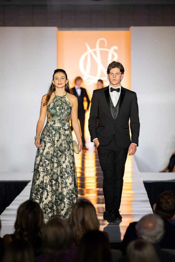 Two models confidently walking down the runway at a fashion show, dressed in elegant evening wear.