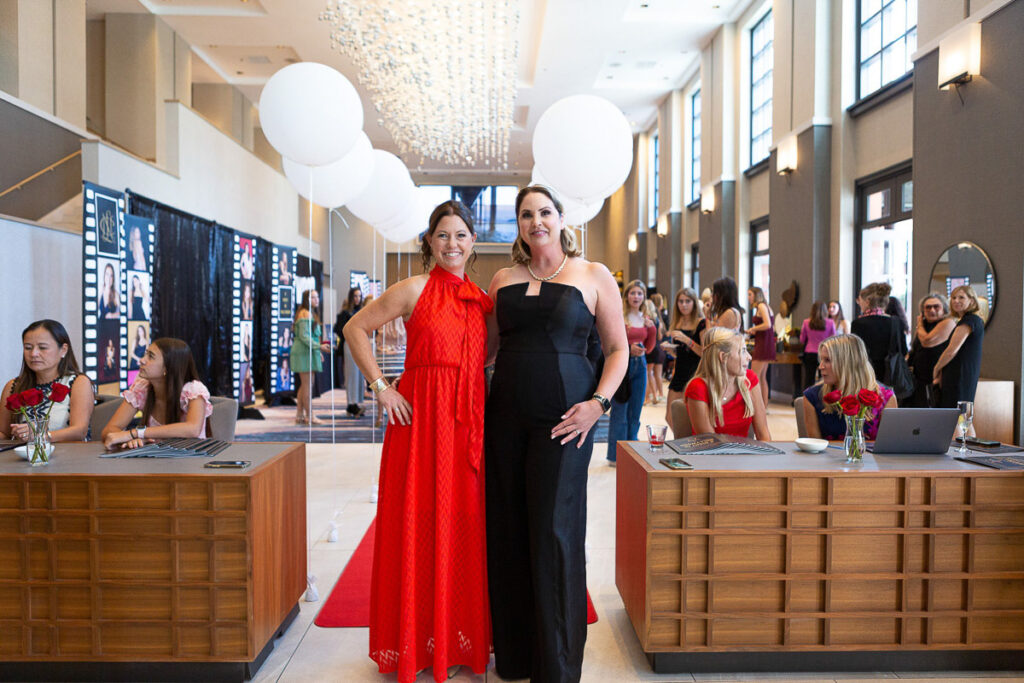 Two women dressed in formal evening wear smiling for the camera at an elegant indoor event with a reception desk and attendees in the background.