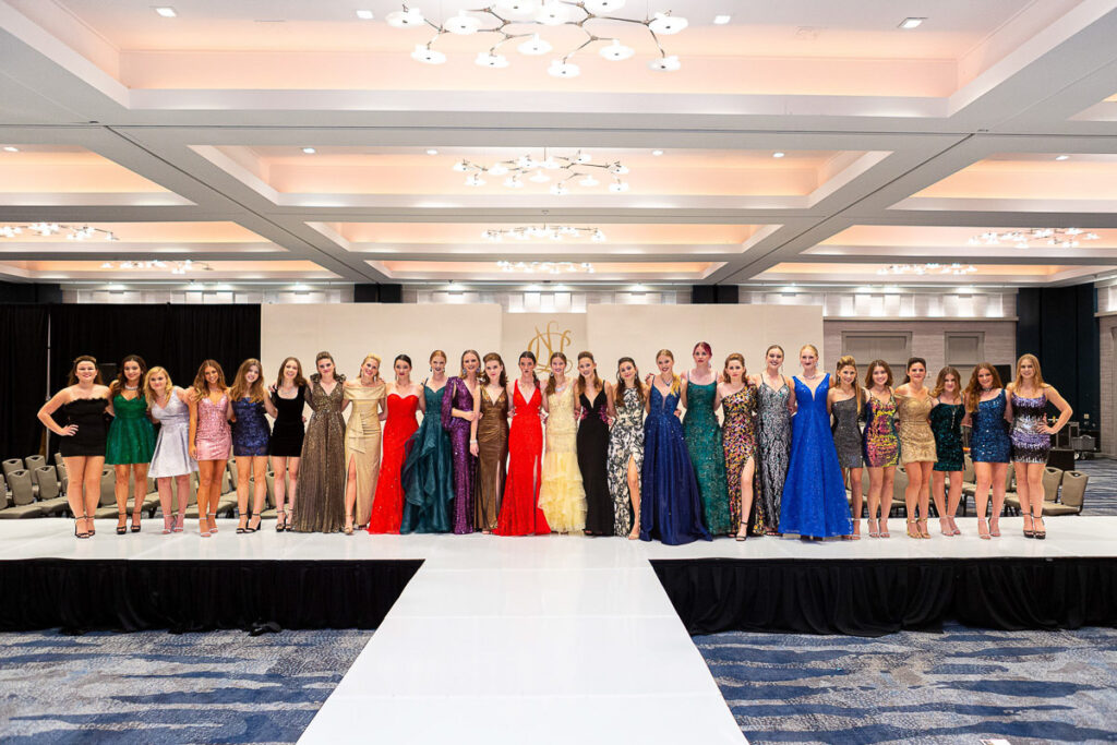 A group of people wearing formal dresses standing confidently in a row on a runway at an elegant event venue.