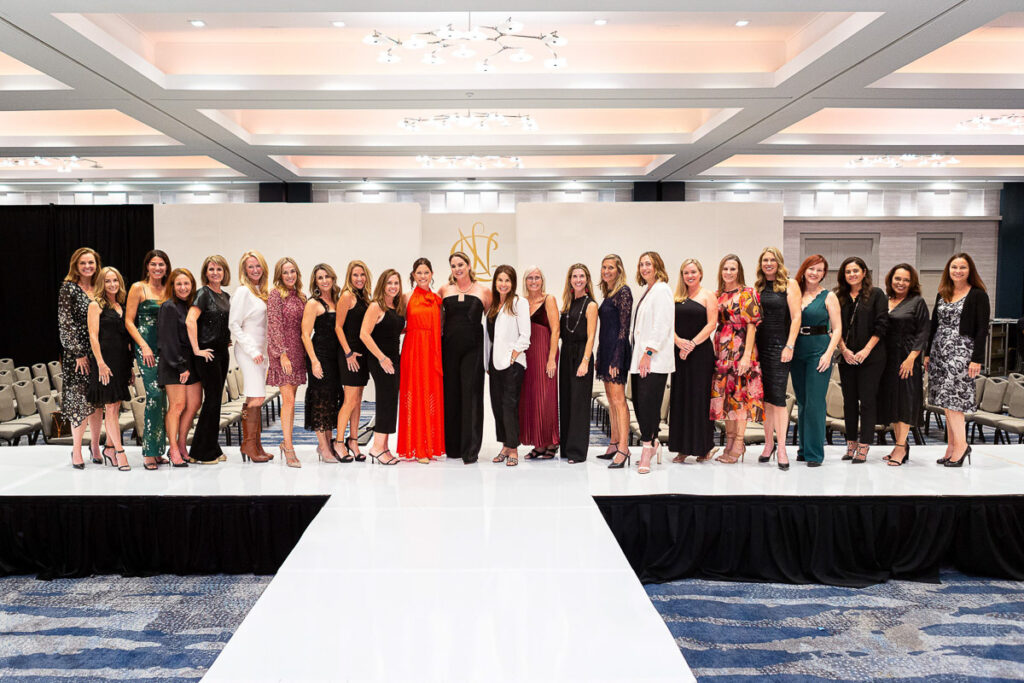 A group of elegantly dressed people posing for a photo on a stage with a blank backdrop and a conference or event setting in the background.