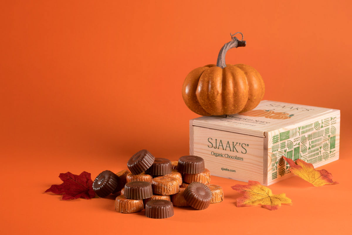 A small pumpkin perched atop an elegant box of Sjaak's organic chocolates, surrounded by an assortment of chocolates and colorful autumn leaves against a warm orange backdrop, evoking a cozy fall atmosphere in