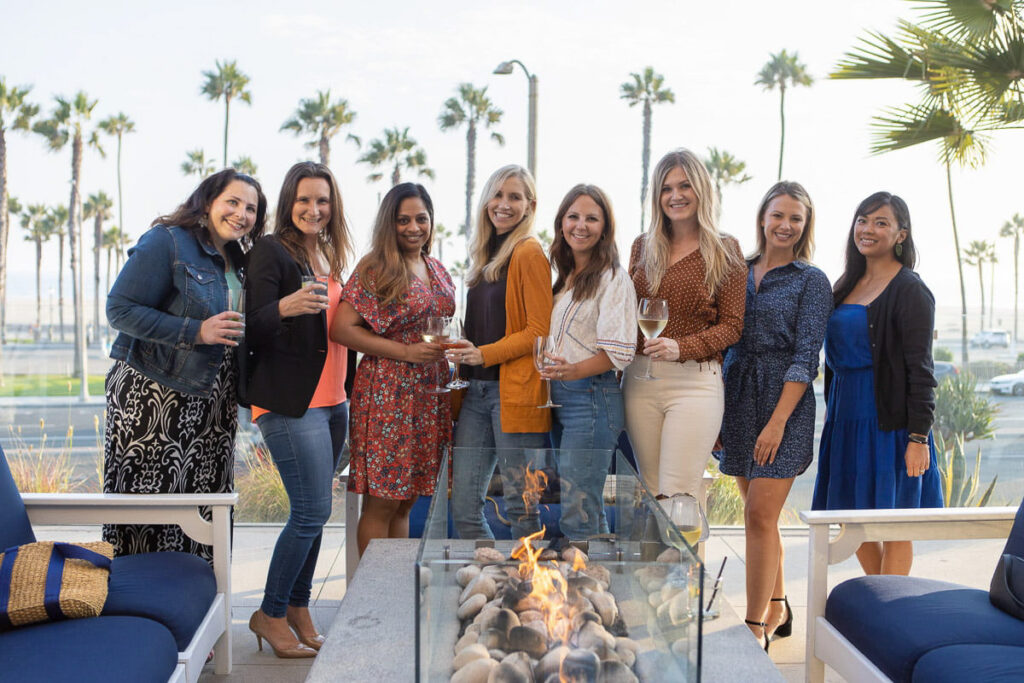 A cheerful group of women enjoying an outdoor gathering by a fire pit with a coastal backdrop, toasting with drinks in hand.