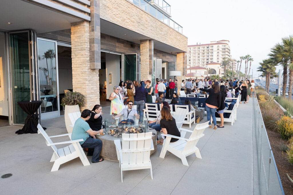 An outdoor social event by a modern building with people mingling and enjoying conversations around fire pit tables.