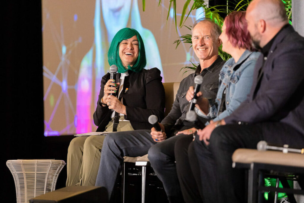 A panel discussion in progress at a conference with a joyful woman speaking into a microphone, sharing a light moment with fellow panelists who are attentively listening and smiling.