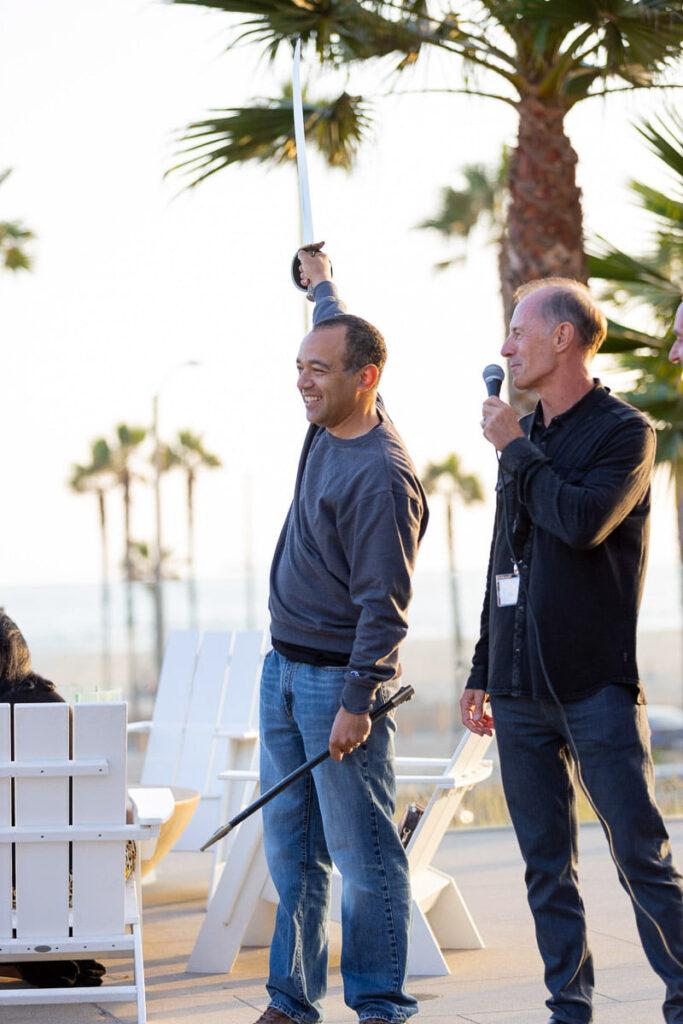 Two men enjoying a lighthearted moment outdoors at a conference, one speaking into a microphone while the other raises his hand with a smile wielding a sword, against a backdrop of palm trees and a setting sun