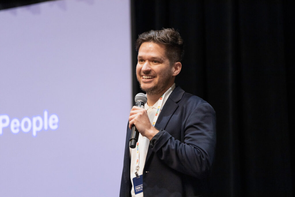 A man smiling while speaking into a microphone at a conference. he stands in front of a screen displaying the word "people". he wears a lanyard and a dark shirt.