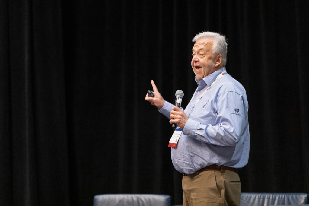 An elderly man speaking passionately at a conference, gesturing with one hand while holding a microphone in the other. he wears a casual shirt and has a conference badge.