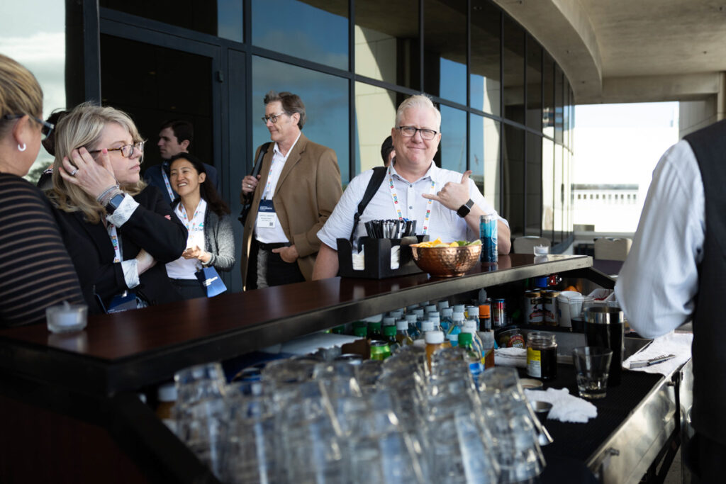 A group of professionals, wearing conference badges, conversing and smiling around a bar table with drinks and snacks, in a modern building with large windows.