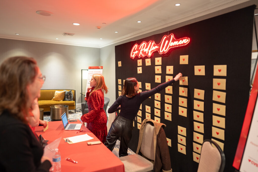 Two women interact at a conference, one pointing at a board labeled "griffin women" adorned with numerous notes. the setting includes a lobby-like area with modern furnishings.