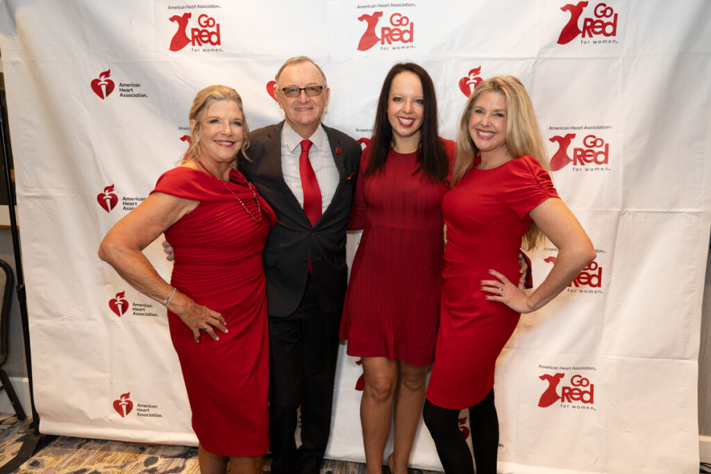 Four people smiling for a photo at a "go red for women" event. two middle-aged women and a younger woman in red dresses flanking an older man in a suit with a red tie, posing in front of a logo backdrop.