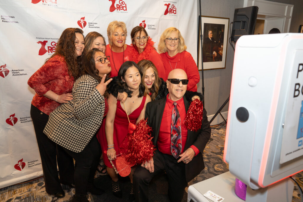 A group of nine people taking a fun photo at a heart association event, dressed in red outfits and accessories, smiling and posing playfully near a photo booth.