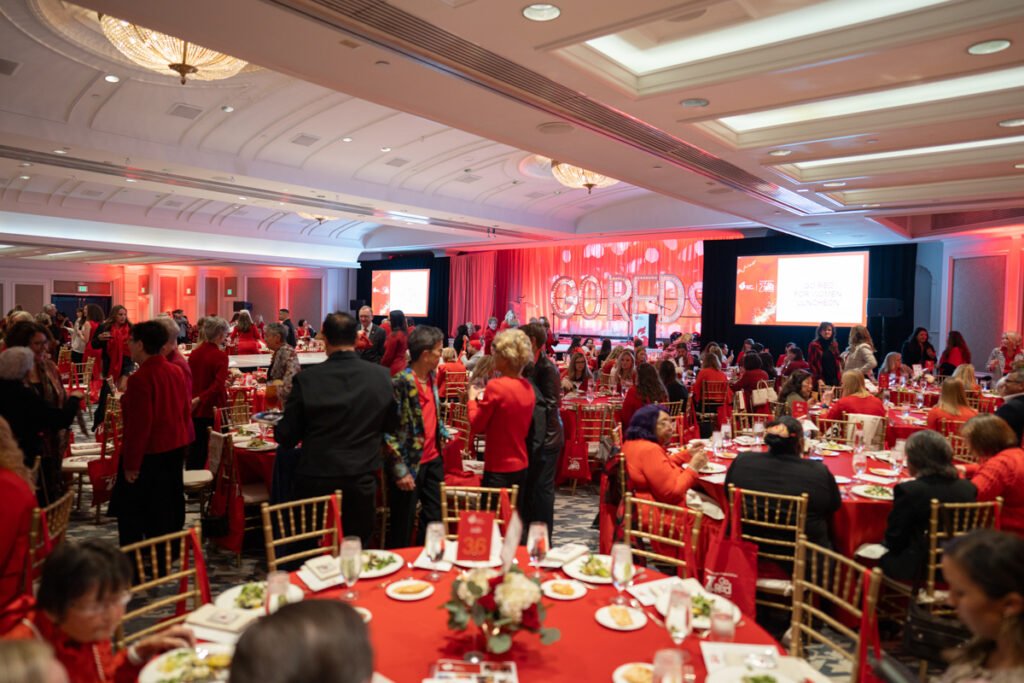 A large banquet room filled with people dressed in red, sitting at tables set for a meal. two large screens display event information. the room is decorated with vibrant red lighting and banners.