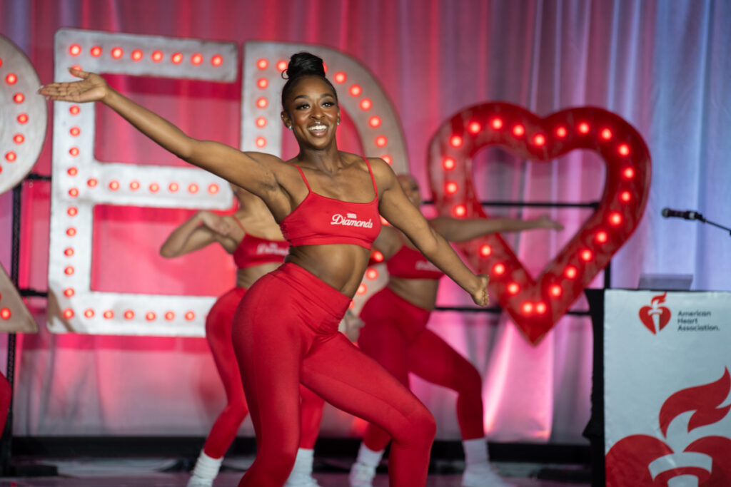 A joyful dancer performs on stage, wearing a red crop top and leggings, against a backdrop with lit-up letters "love" and a heart shape, at an american heart association event.