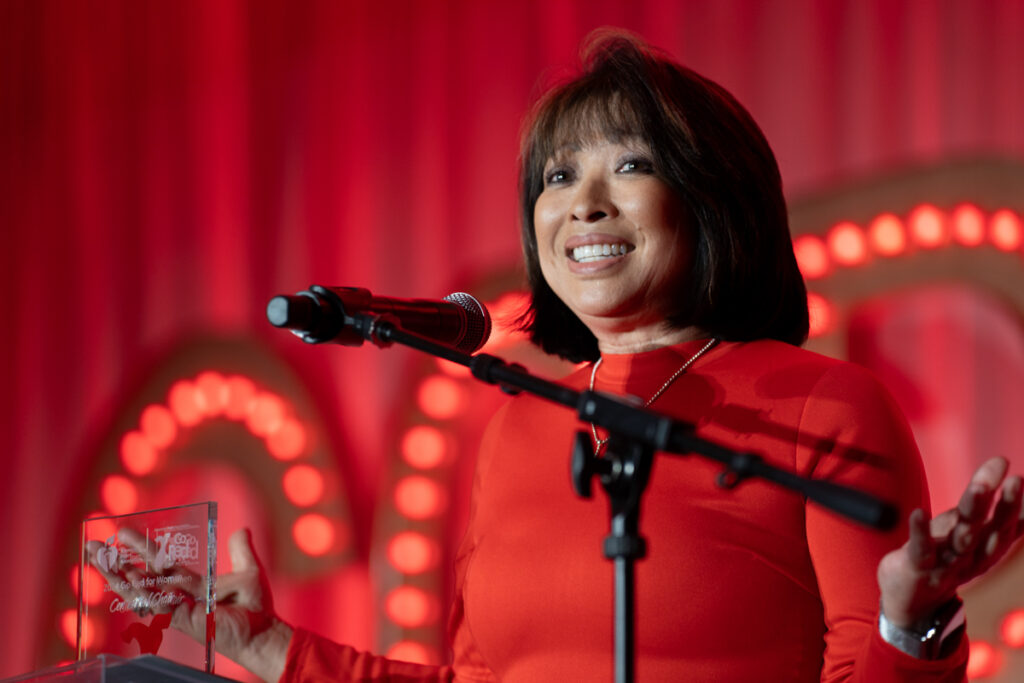 A woman in a red blouse speaking into a microphone at a podium, smiling broadly, with a clear award in front of her and a red, decorated background.