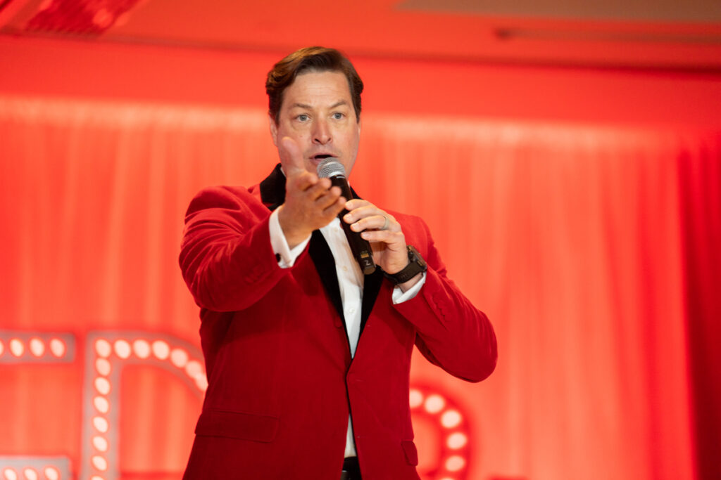 A man in a red blazer and black shirt speaks into a microphone on stage with a tedx sign in the background, engaging an audience with an animated gesture.