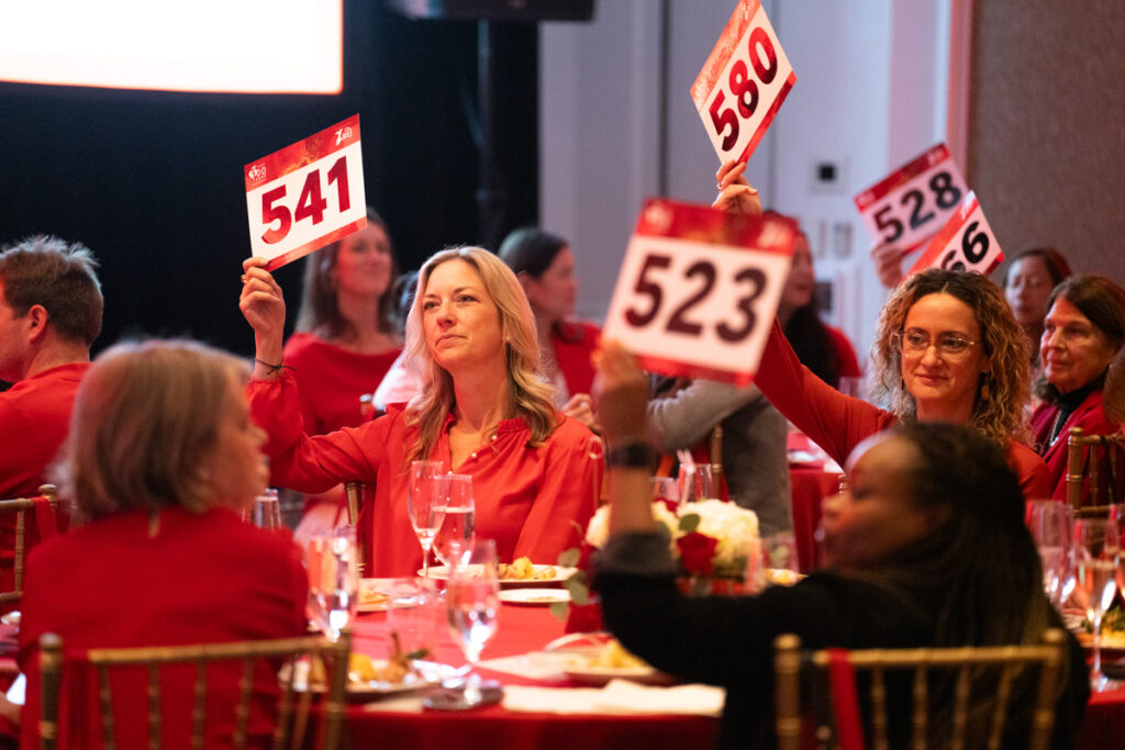 People at a charity auction event, seated at tables and raising numbered red bidding paddles. the focus is on a woman holding paddle number 523. the setting is lively and well-lit.