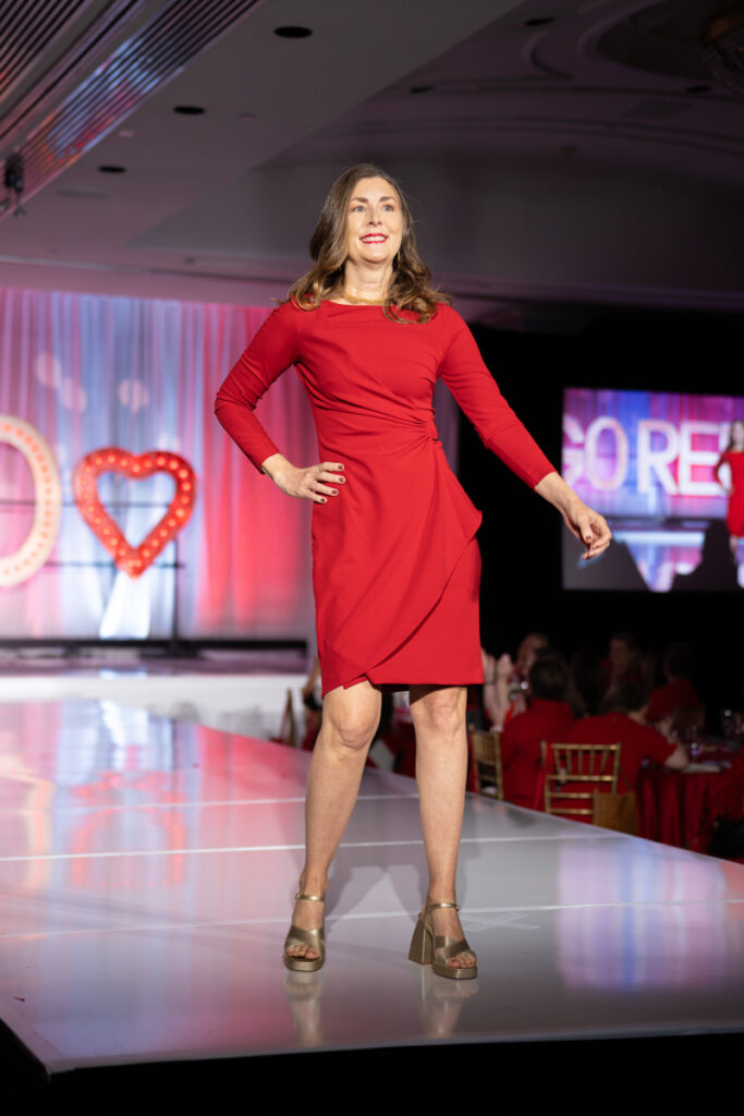 A woman in a red dress confidently walks down a fashion runway, with a bright "go red for women" backdrop, highlighting a heart health event.