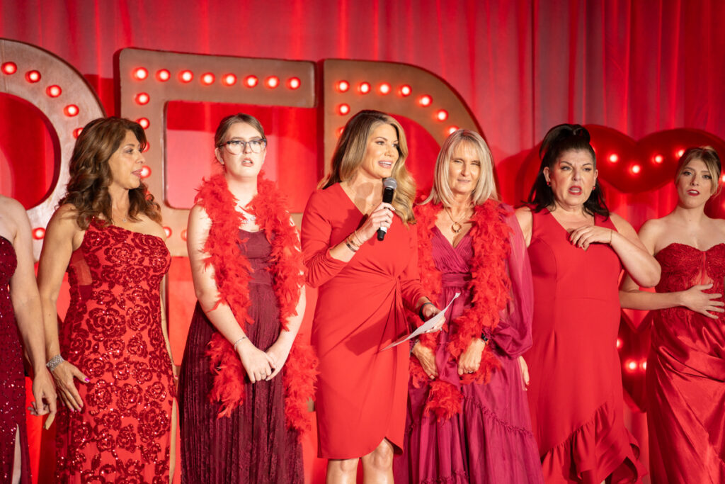 Group of women in red dresses and feather boas on a stage, one holding a microphone, against a backdrop with lighted circles, giving a performance or speech.