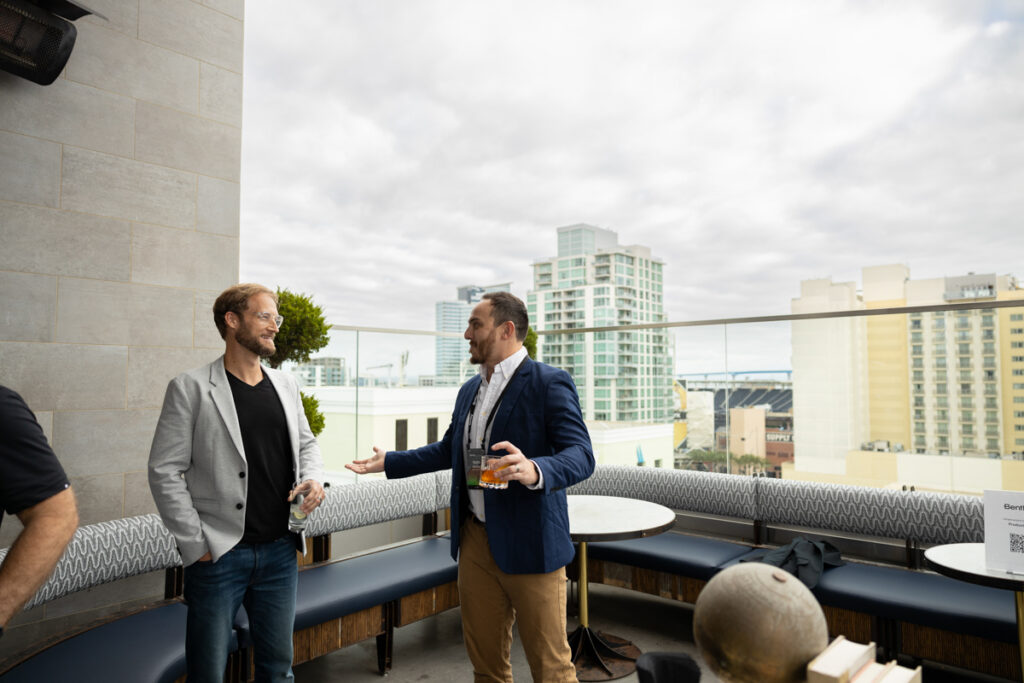 Two men engage in a lively conversation on a rooftop terrace with city buildings in the background. one holds a drink. stylish outdoor furniture surrounds them.