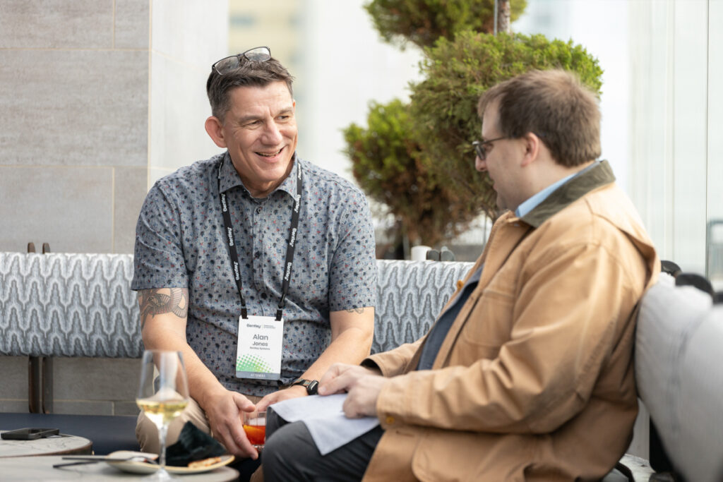 Two men, one holding a drink, engaged in a friendly conversation while seated at an outdoor café, with a plant and a cushioned bench in the background.