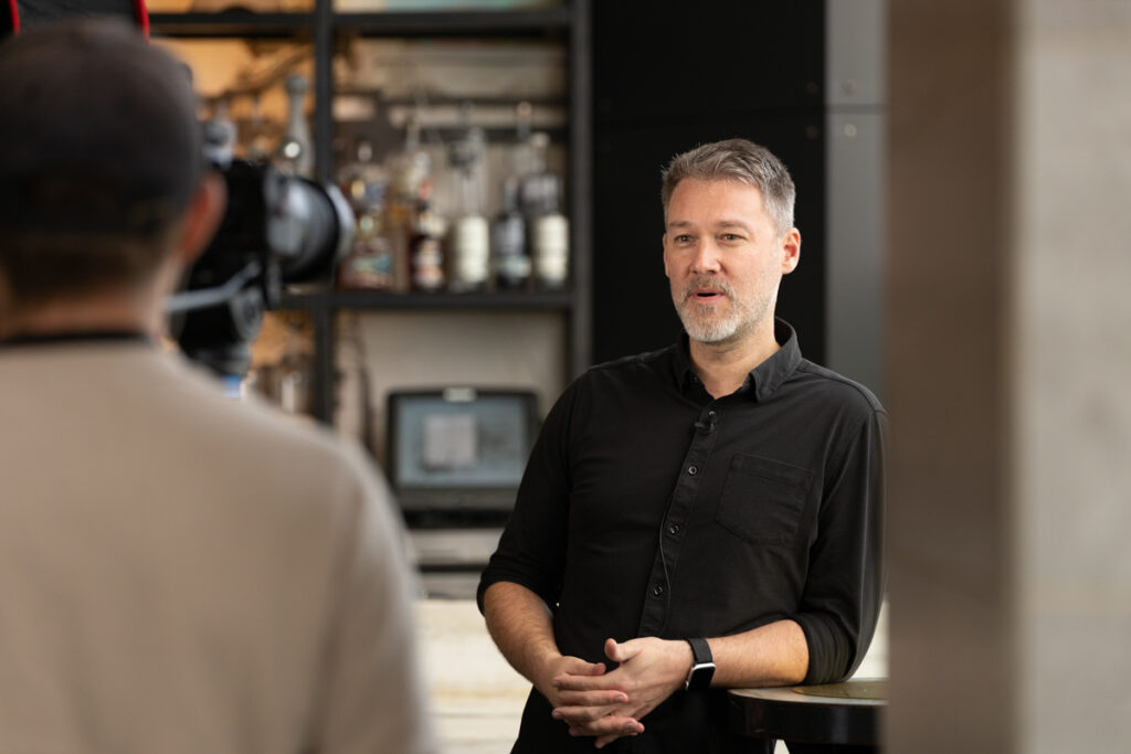 Middle-aged man with graying hair gives an interview, wearing a black shirt, standing indoors in front of a shelf, while a person with a camera records him.