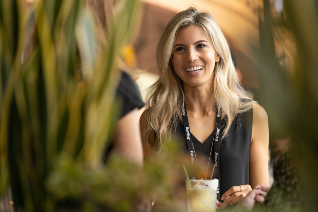 A smiling woman with long blonde hair, wearing a black top and a badge, sitting at a table with a drink in a casual, plant-decorated setting.