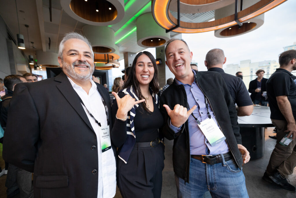 Three professionals at a networking event, smiling and making playful hand gestures in a modern venue with overhead lighting and a crowd in the background.