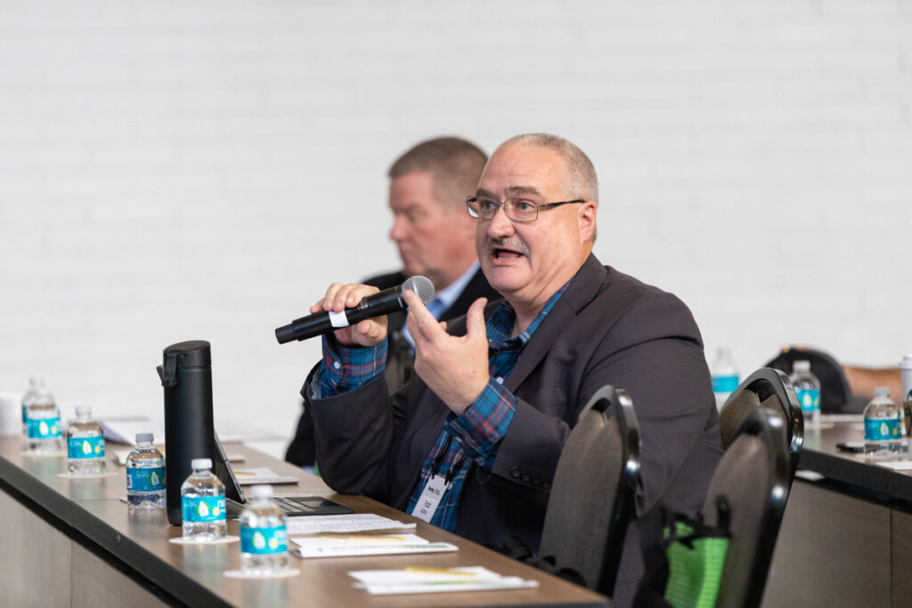 A man wearing glasses and a plaid blazer speaks into a microphone at a conference table, with bottled waters and papers in front, and another participant in the background.