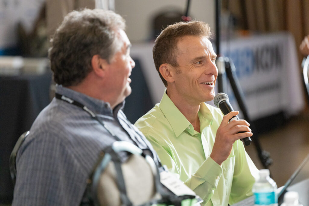 Two men seated at a panel discussion; one speaks into a microphone and smiles while the other, listening intently, looks at him. both appear engaged in a lively conversation.