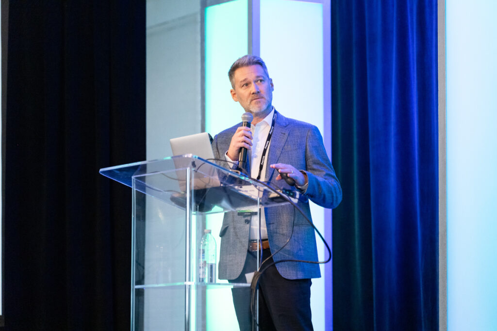 A man in a suit stands at a lectern with a microphone and laptop, addressing an audience at a conference.