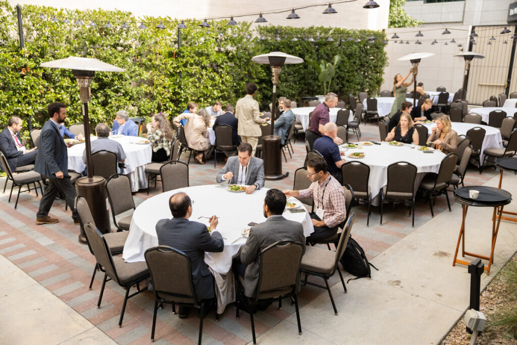 An outdoor networking event with groups of people seated at round tables and eating, surrounded by green hedge walls and patio heaters, with a few individuals standing and conversing.