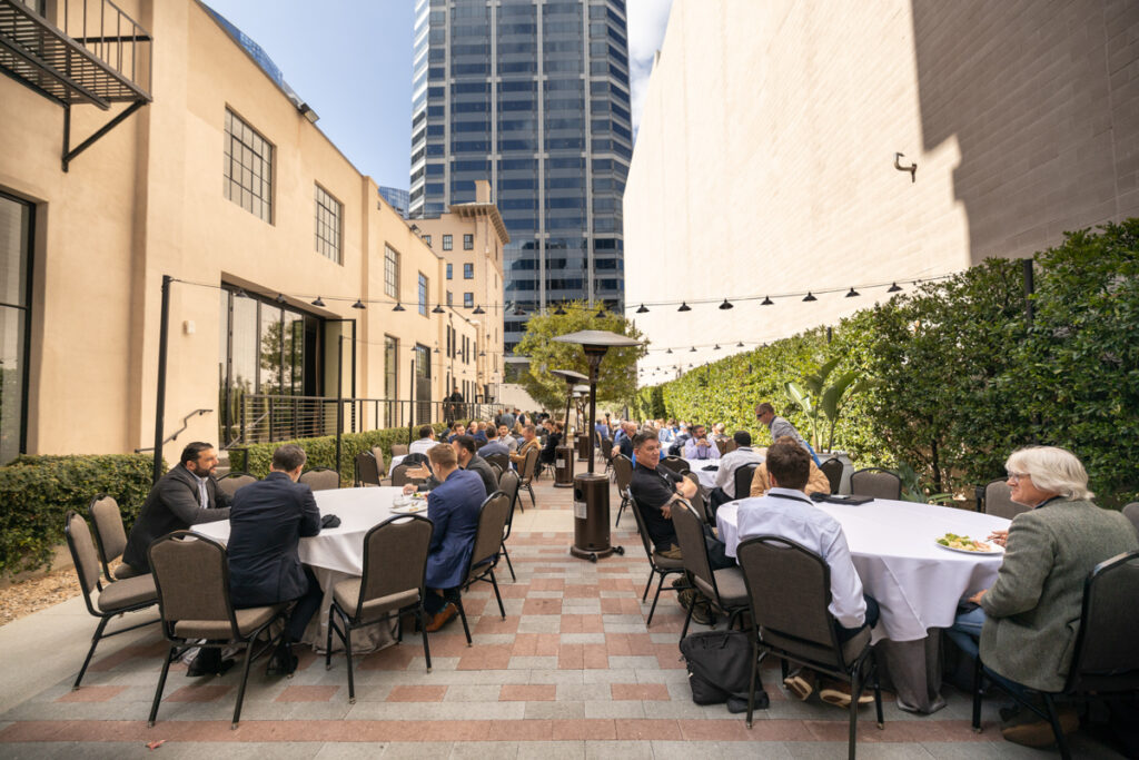 Outdoor corporate event with people seated at tables, eating and conversing, surrounded by buildings, under string lights, with a heater nearby.