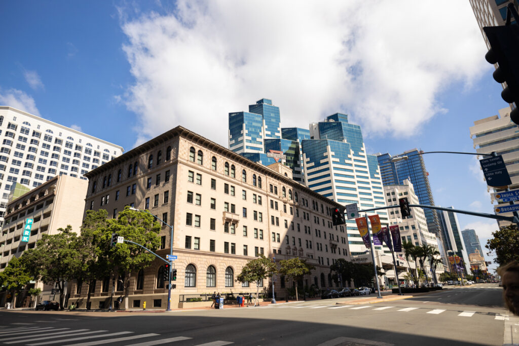 A city street view showing a traditional stone building in the foreground with modern glass skyscrapers in the background, under a clear blue sky.