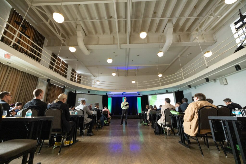 Audience seated at tables listens to a speaker presenting onstage at a conference hall with modern lighting and a balcony.